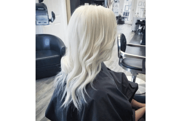 Person with long, wavy platinum blonde hair sitting in a salon chair, facing away.