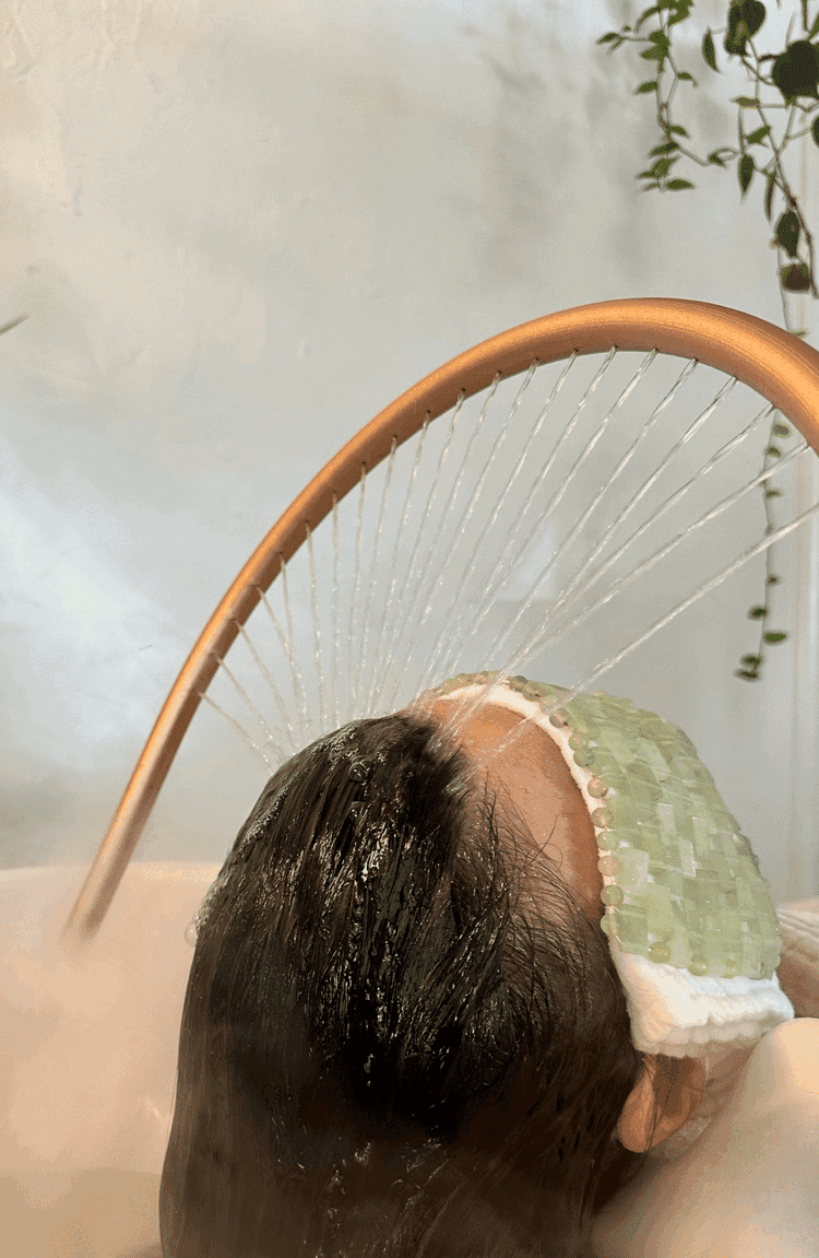 Person relaxing with a green face mask, water spraying over their head in a spa setting.